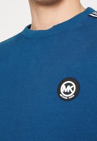 Felpa in cotone blu con colletto a costine e patch circolare nero con le iniziali "MK" e il nome del marchio "MICHAEL KORS."
