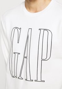 Felpa bianca con colletto a costine e grande testo nero "GAP" stampato in un font alto e stretto sul davanti.