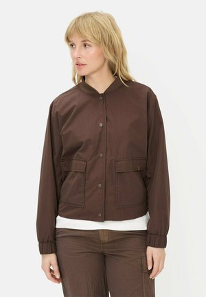 Kobieta o blond włosach, ubrana w brązową zapinaną na guziki kurtkę z klapkowymi kieszeniami oraz pasujące brązowe spodnie, patrząca lekko w lewo.