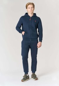 Felpa con cappuccio blu navy con cerniera e tasche a marsupio, abbinata a pantaloni della tuta aderenti coordinati. Presenta un piccolo logo rosso sul petto.