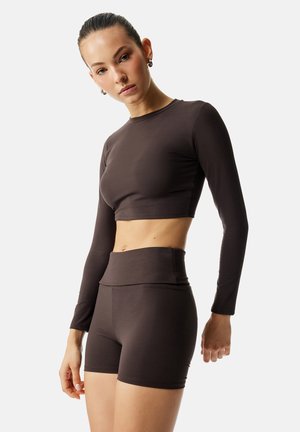 Młoda kobieta z włosami związanymi do tyłu, ubrana w ciemnobrązowy, długi crop top i wysokiej talii krótkie spodenki rowerowe na jednolitym tle.