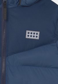 Př paddedná bunda v námořnické modři se zapínáním na zip. Obsahuje logo s šesti kulatými výřezy. Hladká textura s geometrickým vzorem.