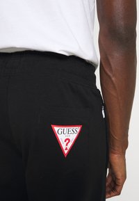 Schwarze Hose mit einer Gesäßtasche, die mit einem roten und weißen Guess-Logo-Patch versehen ist, getragen von einer Person in einem weißen Shirt mit sichtbaren Armvenen.