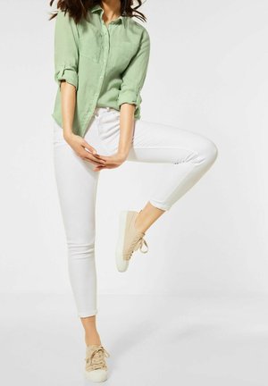 Mujer con camisa verde claro abotonada y jeans blancos, equilibrándose sobre una pierna con zapatillas beige contra un fondo blanco.
