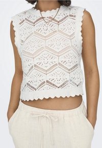Top blanco sin mangas de punto con dobladillo con borde en forma de ondas y un patrón hexagonal intrincado. Se lleva con pantalones claros con cordón.