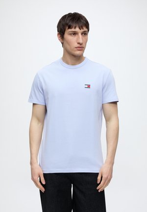 Młody mężczyzna w jasnoniebieskiej koszulce z krótkim rękawem z małym logo Tommy Jeans na piersi, stojący na jednolitym białym tle.