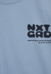 Jasnoniebieska tkanina z czarnym napisem "NXT GAD" oraz mniejszym tekstem "ADAPTIVE WEAR FOR ACTIVE MINDS" i współrzędnymi geograficznymi.