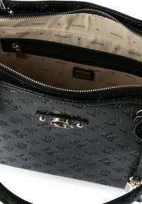 Zwarte Guess-handtas met geperste logo's, zilveren gesp en beige binnenvoering bedrukt met "Guess"-tekst en driehoekpatronen.