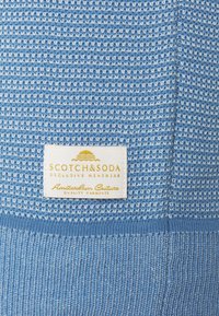 Hellblaues Strickkleidungsstück mit strukturiertem Muster; verfügt am unteren Ende über ein weißes Etikett mit goldener Schrift, auf dem steht "SCOTCH&SODA EXCLUSIVE MENSWEAR".
