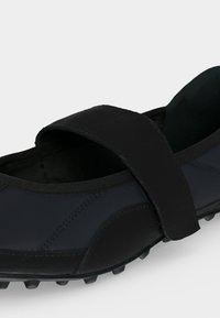 Chaussure Mary Jane noire avec une large bride réglable et une semelle en caoutchouc texturée pour l’adhérence et le confort.