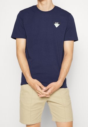 Homme portant un t-shirt bleu marine à manches courtes avec le logo EA7 et un short beige, mains jointes devant, debout devant un fond uni.