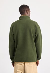 Grøn fleece-pullover med høj krave, der har en tekstureret overflade og orange detaljer i kanten. Designet til varme og komfort.