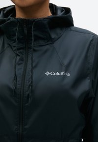 Gros plan sur une personne portant une veste noire Columbia avec une fermeture éclair à l'avant, une capuche et des cordons de serrage.