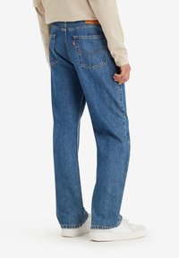 Jean en denim bleu à coupe décontractée, présentant des poches arrière avec des détails de couture et une petite étiquette logo, associé à des baskets blanches.