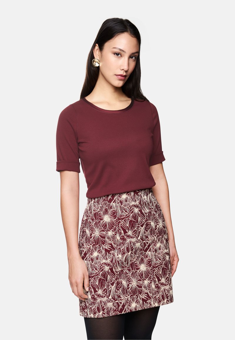 Bordeauxfarbenes Oberteil mit rundem Ausschnitt und kurzen aufgerollten Ärmeln, kombiniert mit einem Rock mit Blumenmuster in ähnlichen Farbtönen und schwarzen Strumpfhosen.