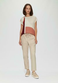 Korte mouwen top met een geometrisch patroon in crème, rood en bordeaux, gecombineerd met beige joggingsbroek met trekkoord en lichtgekleurde platform sneakers.