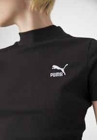 Personne blonde portant un haut noir côtelé à manches courtes avec un col montant et un logo Puma blanc sur la poitrine.