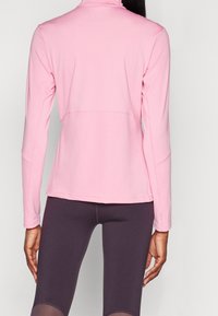 Sportowa bluzka z długim rękawem z miękkiego, jasnoróżowego materiału, z wysokim kołnierzem i dopasowanym krojem, w zestawieniu z ciemnofioletowymi legginsami.