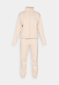 Ensemble de survêtement beige comprenant une veste zippée avec un col montant et un pantalon assorti avec des poignets élastiques. Fabriqué en tissu doux et texturé.