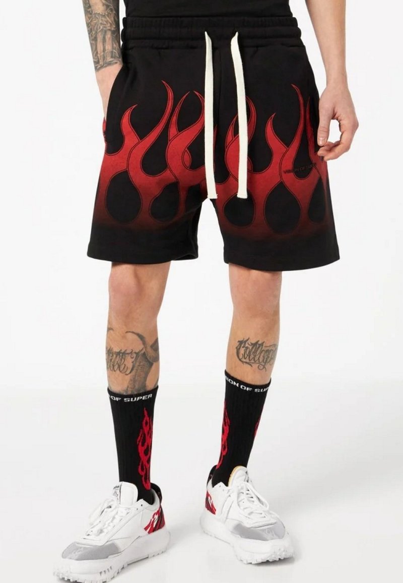 Pantaloni corti neri con motivo di fiamme rosse all'orlo, dotati di cintura con cordino. Indossati con calzini neri e sneaker bianche.