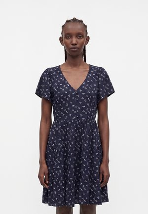 Femme aux cheveux tressés portant une robe bleu marine à manches courtes avec un petit motif floral blanc et une fermeture boutonnée sur le devant.