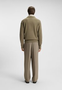 Maglione a maglia verde oliva con colletto sciallato, polsini e orlo a coste, abbinato a pantaloni beige chiaro, visto da dietro. Texture liscia.