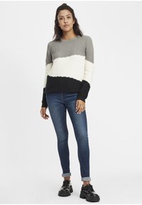 Strikkegenser i grått, svart og kremfarge med ribbestruktur; har et blokk mønster. Kombinert med tettsittende blå jeans og svarte sko.