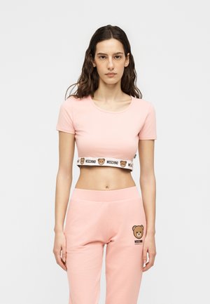 Γυναίκα που φοράει ροζ κοντό τοπ με λωρίδα λογότυπου αρκουδάκι Moschino και ροζ παντελόνι με στάμπα αρκουδάκι στο αριστερό μηρό.