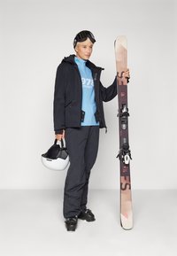 Attrezzatura da sci con una tavola da snowboard di colore marrone chiaro e rosso, utente con giacca e pantaloni neri, maglietta blu, che tiene un casco bianco e sta in piedi.