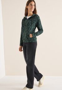Felpa con zip a stampa leopardata verde, dotata di cappuccio con cordino e tasche laterali, abbinata a pantaloni neri e sneakers chiare.
