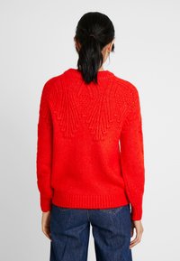 Pull en tricot rouge avec un motif texturé et des poignets et un ourlet côtelés. Présente un design unique au dos. Porté avec un jean bleu.
