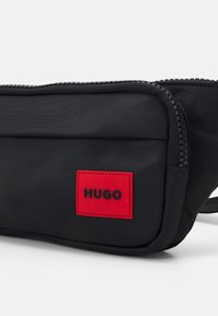Borsa a tracolla nera in nylon con un'etichetta rettangolare rossa e chiusura a zip. Texture liscia con design compatto e tracolla regolabile.