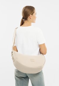 Bežová popruhová taška s volným strihom vyrobená z mäkkej tkaniny s plochým dnom, hrubým popruhom a malým logom značky. Nosená cez telo.