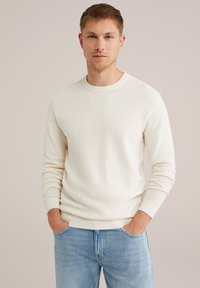 Hellbeige strukturierter Pullover mit gerippten Bündchen und Ausschnitt, kombiniert mit hellblauen Jeans. Der Pullover hat ein dezentes Muster.