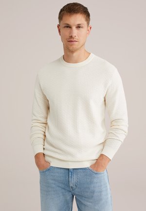 Suéter texturizado beige claro con puños y cuello de canalé, combinado con jeans de color azul claro. El suéter presenta un patrón sutil en toda su superficie.