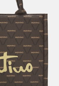 Brun, texturerad läderhandväska med guldtonad "Valentino"-logotyp och upprepande "V"-mönster. Har ett enda handtag.