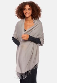 Châle gris clair avec des franges, drapé sur un haut noir. Texture douce et forme rectangulaire, offrant chaleur et couverture.