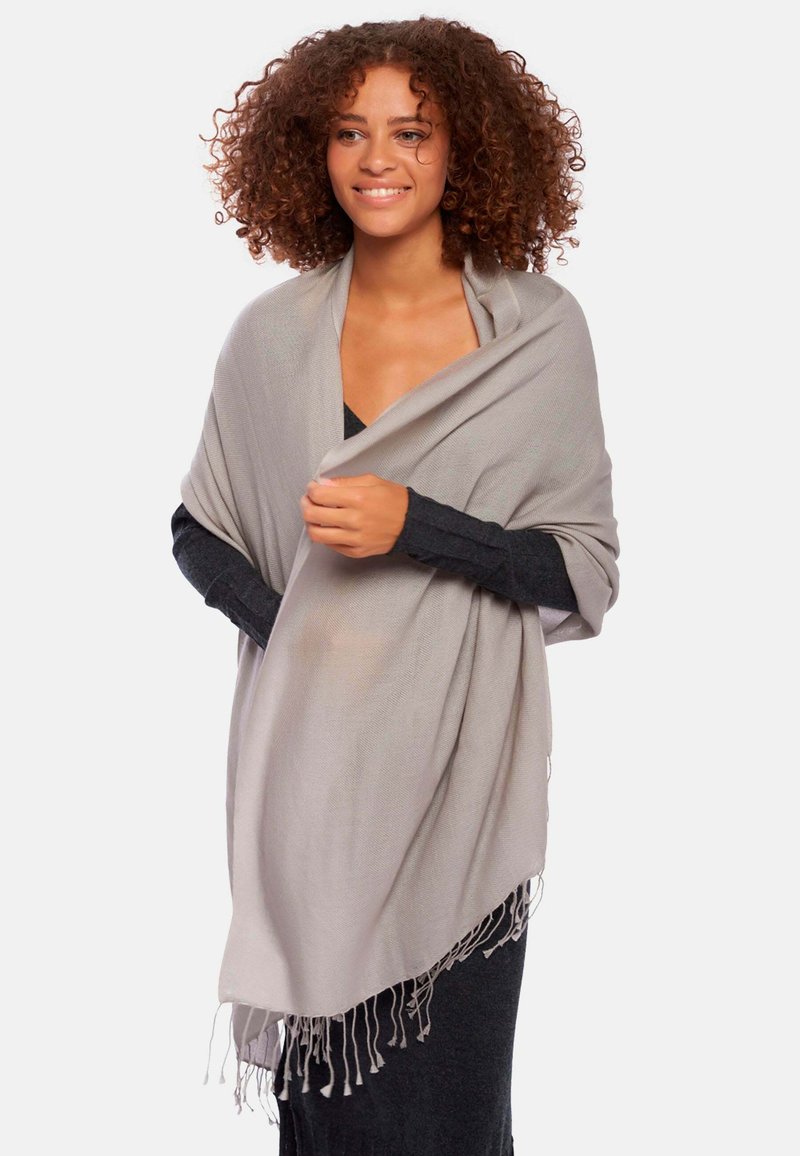 Châle gris clair avec des franges, drapé sur un haut noir. Texture douce et forme rectangulaire, offrant chaleur et couverture.