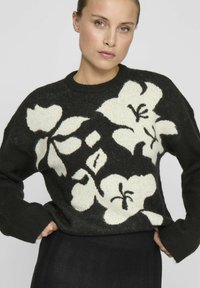 Černý pletený svetr s velkým bílým květinovým vzorem. Dlouhé rukávy a kruhový výstřih. Měkká textura s žebrovanými manžetami.