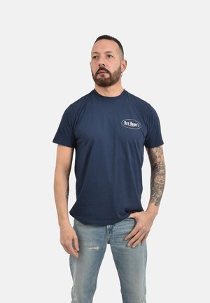 Uomo con barba e braccio tatuato che indossa una maglietta blu navy "Roy Roger's" e jeans blu slavati su sfondo bianco uniforme.
