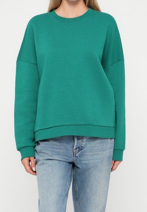 Person trägt grünes Sweatshirt mit Rundhalsausschnitt und abgefallenen Schultern sowie hellblaue Jeans, steht vor einfachem Hintergrund.