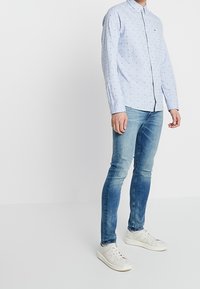 Lichtblauw overhemd met lange mouwen en kleine marineblauwe stippen, met een geknoopte kraag, gedragen met vervaagde blauwe spijkerbroek en witte sneakers.