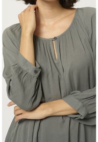 Femme portant un chemisier gris texturé à manches longues avec un décolleté en forme de trou de serrure et des boutons, bras croisés avec une main près du cou.