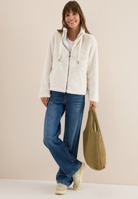 Veste blanche duveteuse zippée avec capuche, cordons de serrage et poches latérales, associée à un jean bleu et un sac vert, portée avec des baskets beiges.