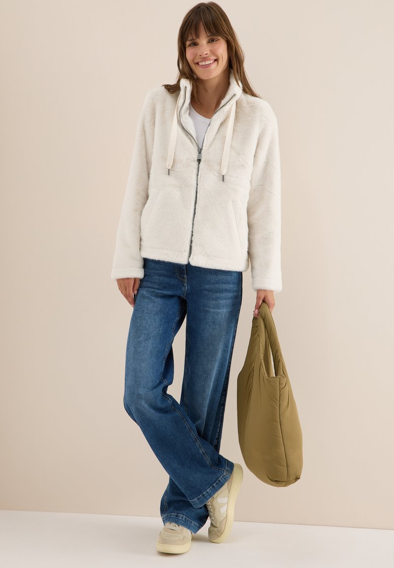 Veste blanche duveteuse zippée avec capuche, cordons de serrage et poches latérales, associée à un jean bleu et un sac vert, portée avec des baskets beiges.