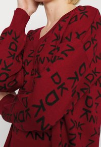 Roter gestrickter Pullover mit tiefem V-Ausschnitt, der ein sich wiederholendes Muster aus schwarzen Buchstaben aufweist. Die Ärmel sind leicht übergroß.