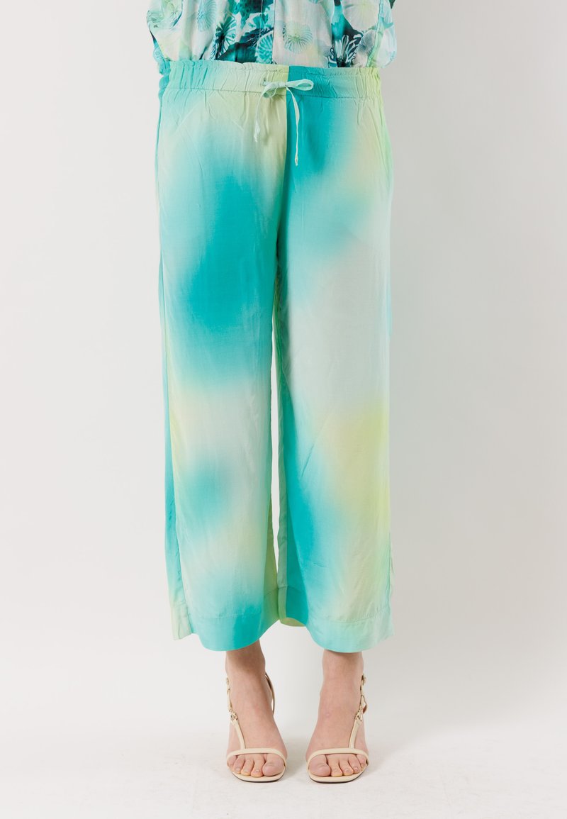 Donna che indossa pantaloni larghi e ampi con motivo tie-dye blu, verde e bianco, abbinati a sandali beige aperti in punta.