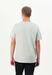 Mand med kort, krøllet hår iført en ensfarvet, lysegrå t-shirt med korte ærmer og sorte bukser, som vender væk på en hvid baggrund.