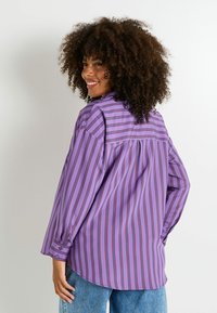 Camisa de manga larga en tela morada con rayas rojas verticales, que presenta un puño abotonado y un detalle de yugo plisado en la espalda.