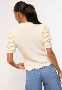 Svetrobéžový pletený sveter s textúrovanými nafúknutými rukávmi, zdobený vlnitými okrajmi a jemnými čipkovými detailmi na leme a rukávoch.